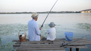 loving father, caring parent spends time with his child talking to him while sitting on pier near river and fishing on fishing rod during summer holidays